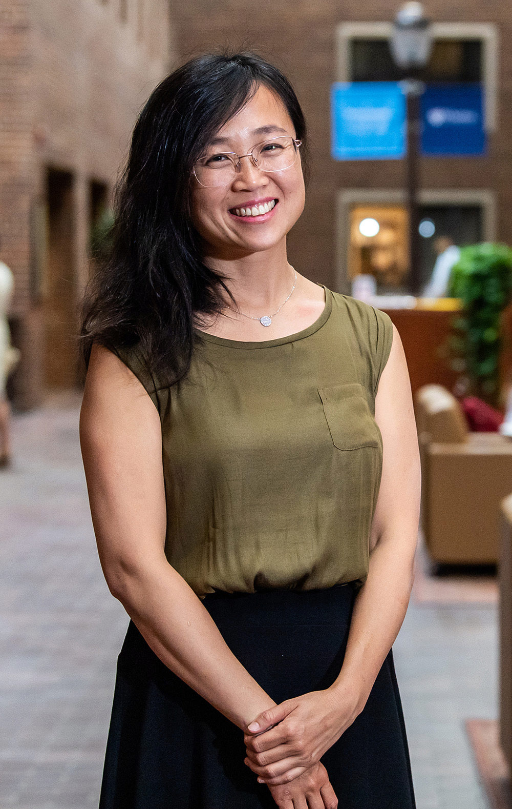 A person smiling indoors, wearing glasses and a green sleeveless top, standing in a brick-walled environment with signs and furniture in the background.