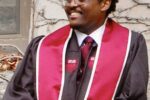 A person wearing a graduation gown and red stole, smiling while standing in front of a wall with climbing vines.