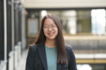 A person with long hair and glasses is smiling, wearing a blazer and a teal top, standing in a hallway with a blurred background.