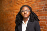 Headshot of a person with glasses and curly hair, wearing a white shirt and dark blazer, standing in front of a brick wall.