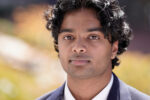 A professional headshot of a person wearing a dark suit and white shirt, with a neutral expression and a blurred outdoor background.