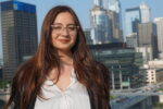 A person with long brown hair and glasses, smiling, is in front of a cityscape featuring tall buildings and a clear blue sky.