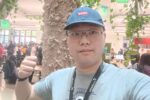 A person wearing glasses and a blue cap gives a thumbs-up in a bustling indoor setting with people working and socializing. There is a tree trunk and green decor in the background.