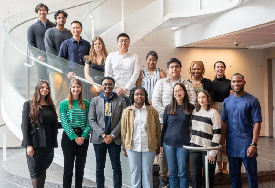 A diverse group of people standing together indoors in front of a modern spiral staircase, appearing to be a professional or academic group portrait.